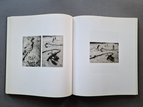 An open book showing two black-and-white photographic prints on the left page and one larger black-and-white print on the right page, photographed from above on a dark gray surface.