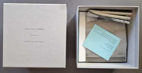 An open box displayed next to its lid on a gray surface. Inside the box are folded neutral-toned paper items and a light blue card with printed text.
