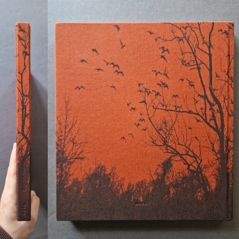 A square book with a textured red background and dark almost black silhouetted trees and birds, shown from the front and side. A hand is holding the spine on the left against a gray background.