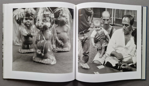 An open book showing two black-and-white photographs side by side. The left photo shows several small plastic-wrapped figurines or dolls on the floor.