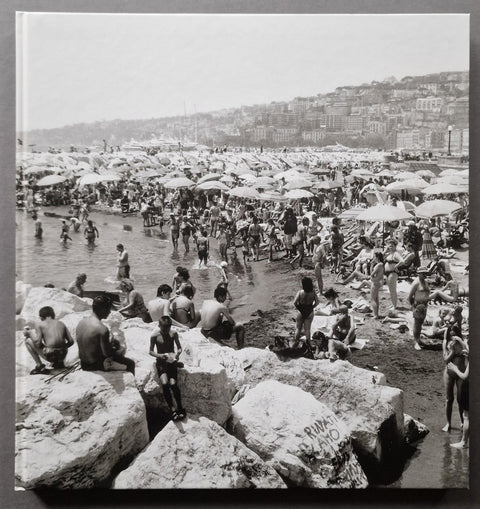 A black and white square cover of a crowded beach scene with people swimming, sitting on large rocks, and umbrellas scattered across the shore, with a hillside town in the background.
