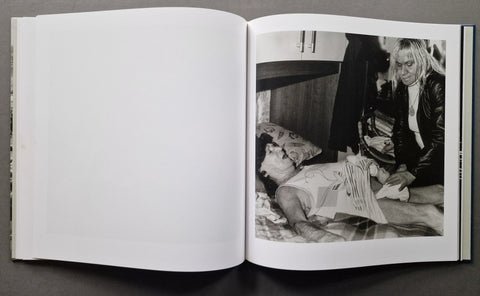 An open hardcover book laid flat on a dark surface, showing a black-and-white interior photograph on the right page with a person lying down and another person standing nearby, while the left page is blank.