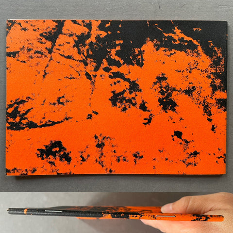 An orange and black abstract patterned notebook or journal is shown standing upright on a gray surface, with a hand holding a matching slim notebook at the left edge.