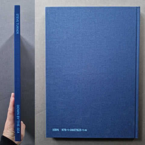 A hardcover book titled 'Down by the Sea' by Steve Turner, shown standing upright next to its spine, with a hand holding the spine for scale. The book has a textured blue cloth cover with light blue text.
