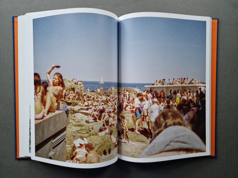 An open hardcover book spread, showing two printed pages with a large vintage beach photograph. The photo depicts a crowded beach scene with people, rocks, blue sky, and ocean.