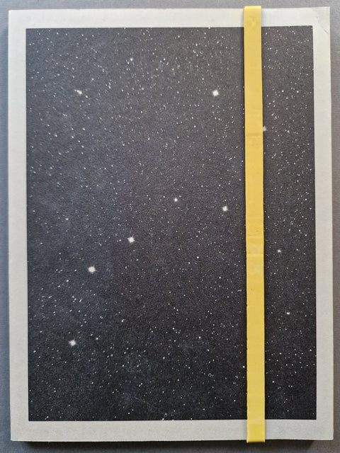 A paperback book cover with a dark starry sky cover, featuring a vertical yellow elastic closure band and a grey border.