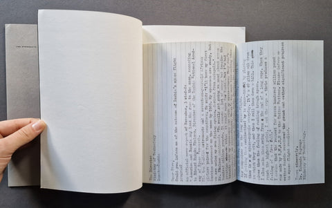 An open book lying on a dark surface, with a hand holding the left page open. The left page is blank, while the right pages contain typed text in black ink on white paper.