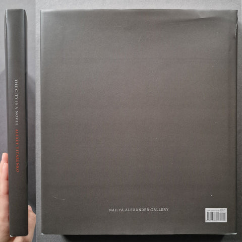 A hardcover book or art print shown from the back, with a hand holding the spine on the left. The cover is a dark charcoal gray with small white text reading 'NAILYA ALEXANDER GALLERY' and a barcode in the lower right.