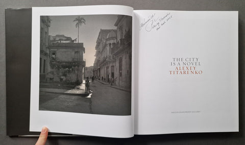 An open hardcover book or art print showing a black-and-white street photograph on the left page and a white text page on the right with a signature in the corner.