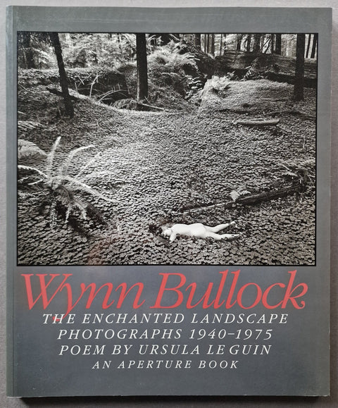 Book cover of 'Wynn Bullock: The Enchanted Landscape' featuring a black and white photograph of a forest path with a body on the ground, and red and white text on a gray background.