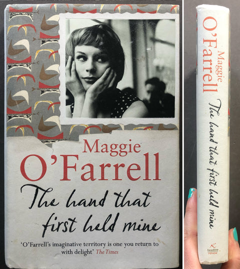 A book titled "The hand that first held mine" by Maggie O’Farrell is shown from the front cover and spine. A hand with teal nail polish holds the spine on the right side.