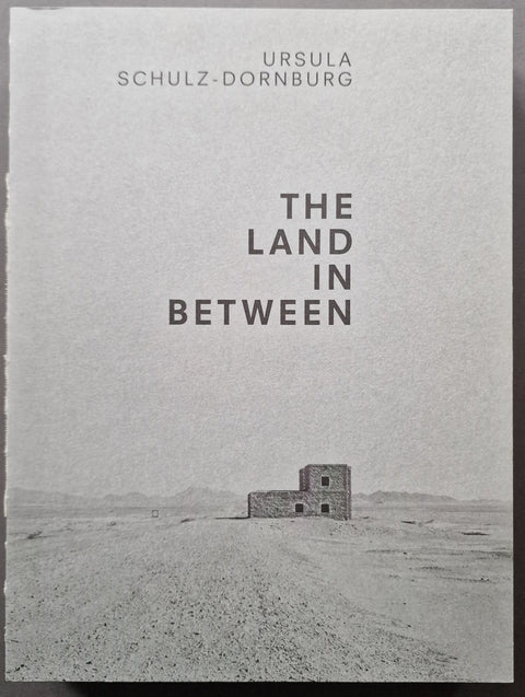 Book cover of 'The Land in Between' by Ursula Schultz-Dornburg, featuring a minimalist black and white photograph of a small stone structure in a vast desert landscape.