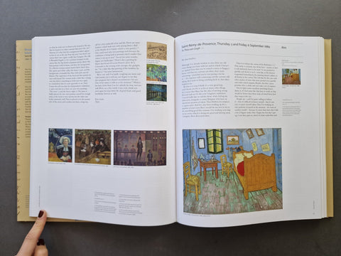 An open book spread showing printed text and multiple paintings, photographed from above on a dark gray surface. A hand with dark nail polish is visible holding the left page edge.