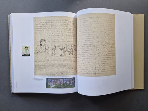 An open hardcover book laid flat on a dark gray surface, showing two spread pages with handwritten text, a sepia-toned illustration, a small inset print, and a landscape painting near the bottom.