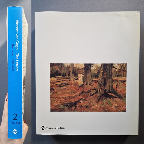 A book titled 'Vincent van Gogh - The letters' shown standing upright next to its cover, held by a hand on the left. The spine is bright blue with white text, and the cover is white with a central painting of a forest scene and the 'Thames & Hudson' logo at the bottom.