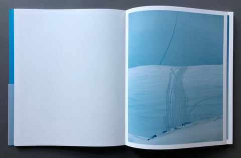 An open spread of to-skip-a-sinking-stone by daniel-dorsa laid flat on a dark gray surface, showing a large blank white page on the left and a photographic print of a snowy blue landscape on the right.