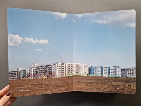AN open book showing a construction site with a dirt foreground, a row of apartment buildings in the distance, and a blue sky with clouds.