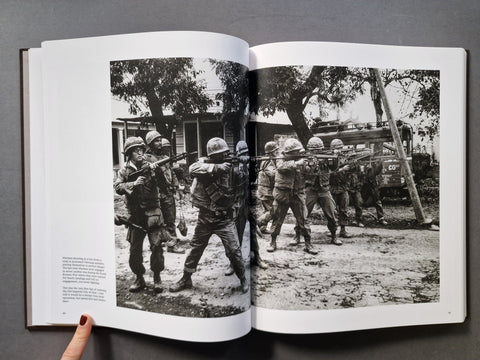 An open book showing a black-and-white historical photograph print of soldiers in motion, held open by a hand against a dark gray background.