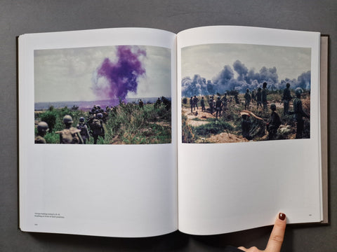 An open book displaying two photographic prints side by side. The left print shows soldiers in a field with a dramatic purple smoke plume in the distance, while the right print shows soldiers standing in a dusty landscape under dark smoke.