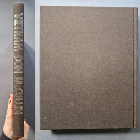 A hardcover book titled 'Vietnam: Don McCullin' is shown standing upright next to a hand holding its spine. The cover and spine are a textured taupe-brown fabric, with the title and author name raised in a lighter metallic or beige tone.