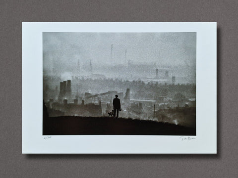 A framed or matted art print showing a misty industrial cityscape with a small dark silhouette of a person standing on a hill overlooking the skyline. The print is photographed against a plain taupe-gray background.