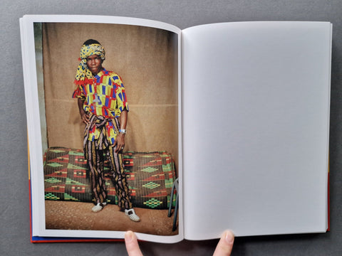 An open book or magazine held at the bottom, showing a printed fashion photograph on the left page of a person wearing a colorful patterned shirt and striped pants, with an empty white page on the right, against a dark gray background.