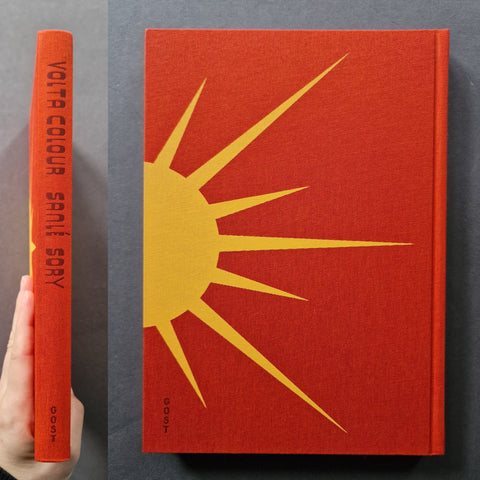 A hardcover book titled 'Volta COLOUR Sanle Sory' shown standing upright next to a hand holding its spine. The cover and spine are a textured red-orange with a large yellow sunburst graphic.