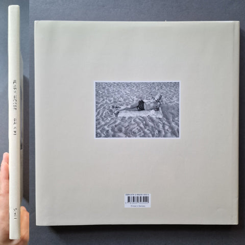 A square art book, featuring a central black-and-white photograph of a person lying on sand.