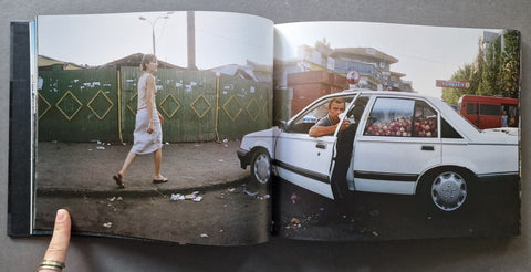 An open book showing a printed photograph of a woman walking beside a white car on a street, held open by a hand of a man. Car is full with apples.
