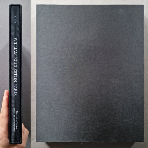 A large black hardcover book or art print shown from above, with its spine visible on the left and held by a hand. The spine reads 'WILLIAM EGGLESTON PARIS' with smaller text below, and the cover is a matte black textured material.