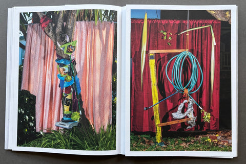 An open art book or photo journal laid flat on a dark surface, showing two printed photographs side by side. The left page features a colorful outdoor scene with pink curtains and a painted tree trunk; the right page shows a red curtain backdrop with hanging blue hoses and other industrial-style objects.