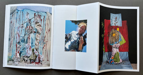An open art book or magazine laid flat on a dark surface, showing three printed pages with colorful artwork: a left page with a blue-toned abstract scene with red drips and figures, a center page with a small portrait photo, and a right page with a surreal painted scene featuring a figure in front of a red backdrop.