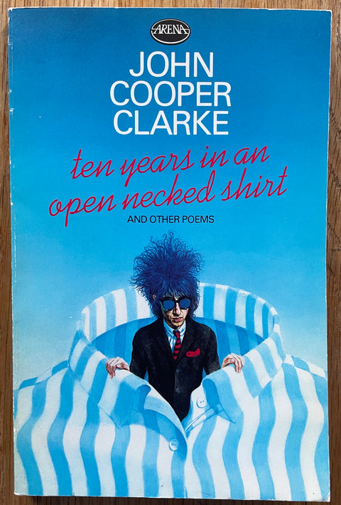 The book cover of Ten Years in an Open Necked Shirt by John Cooper Clark. Softcover in blue with a blue haired man coming out of the collar of a shirt.