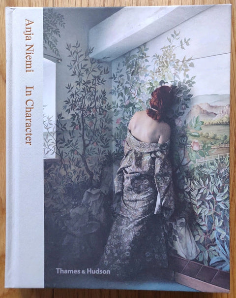 The photography book cover of In Character by Anja Niemi. Hardback with a woman standing with her head on a leafy wallpaper-covered wall.