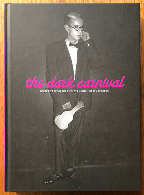 The photography book cover of The Dark Carnival by Derek Ridgers. Hardback in black with pink text for the title.