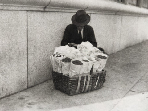 Black and white photograph of a man in a dark suit and hat sitting behind a large woven basket filled with wrapped items on a sidewalk beside a stone wall.
