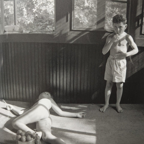 Black and white vintage photograph showing a young boy standing in shorts near a window while a woman reclines on the floor nearby.