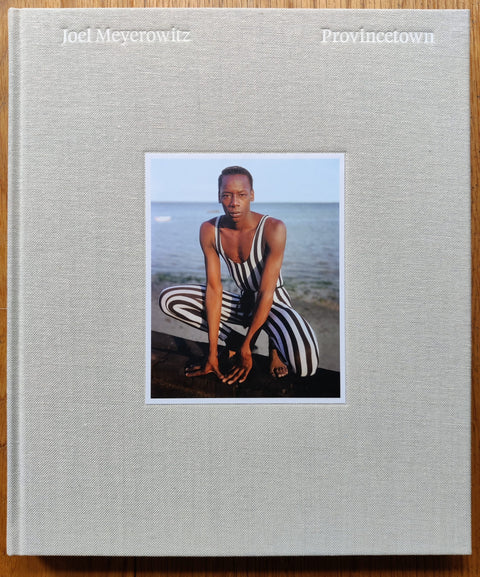 The photography book cover of Provincetown by Joel Meyerowitz. Hardback in grey with a man squatting by the sea in a stripey jumpsuit.