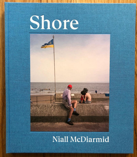 The photography book cover of Shore by Niall McDiarmid. Hardback in blue with photograph of some people sitting on a ledge by the sea.