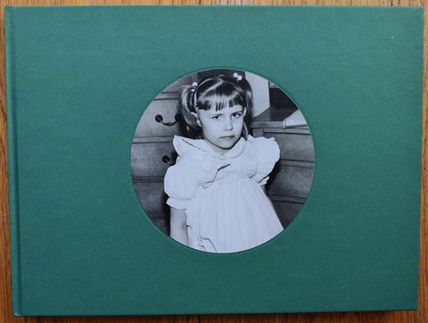 The photobook cover of At Home by Susan Kandel. Hardback in dark green with b&w image of a small girl looking angrily at the camera.