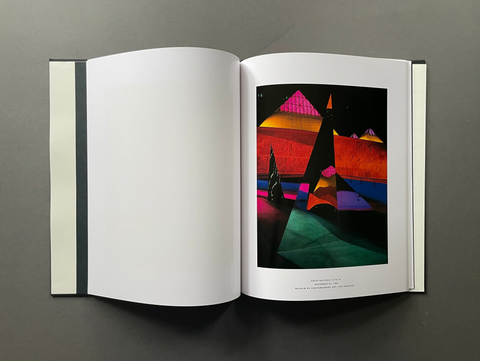 An open art book displayed flat on a dark gray surface, showing a blank white left page and a colorful abstract geometric print on the right page.