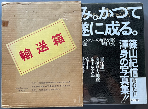 A vintage Japanese shipping box in brown cardboard with a yellow label and red characters, placed next to a black book or booklet with white and Japanese text, on a dark blue-gray background.
