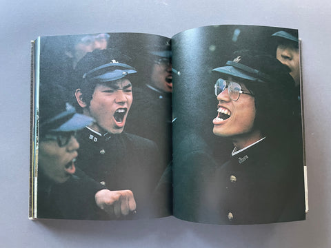 An open book or photo album showing a printed photograph of several people in dark uniforms, with one person shouting and another wearing glasses and a cap, photographed against a plain gray background.