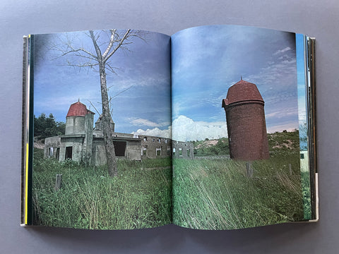 An open hardcover book laid flat on a gray surface, showing a large photographic print spread across both pages. The photo depicts an outdoor scene with tall grass, a leafless tree, and two brick structures under a blue sky.