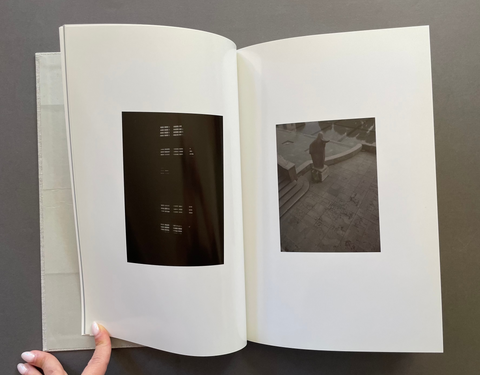 An open white photo book held by a hand, showing two black-and-white photographic prints on the pages against a dark gray background.