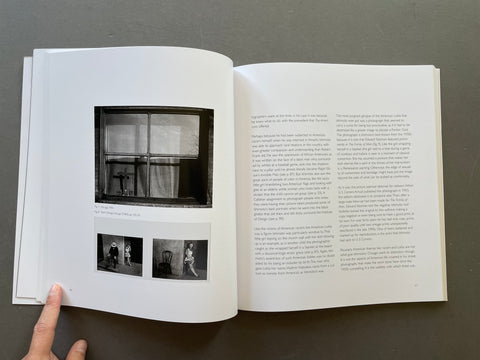 An open book or art print spread on a dark gray surface, showing a black-and-white photograph of a window and smaller black-and-white figures on the left page, with dense text on the right page. A hand is holding the left edge of the book.