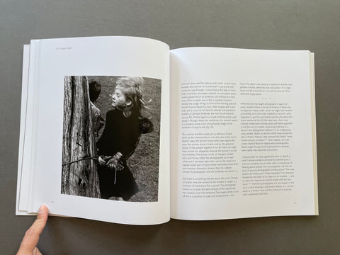 An open book showing a black-and-white photograph of a woman leaning against a tree on one page and columns of text on the other, held open by a hand against a dark gray background.