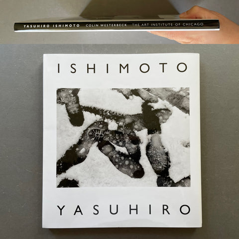 A hardcover art book titled 'ISHIMOTO YASUHIRO' is held by a hand above its cover, which features a black and white abstract photograph. The book is photographed on a gray surface.