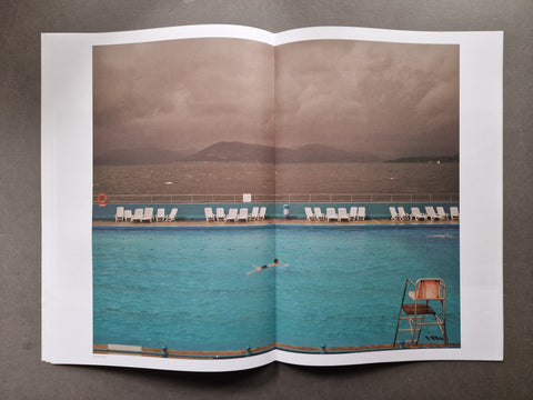 An open publication spread photographed from above on a dark gray surface, featuring a large photographic print of a swimming pool with lounge chairs, distant hills, and a cloudy sky.