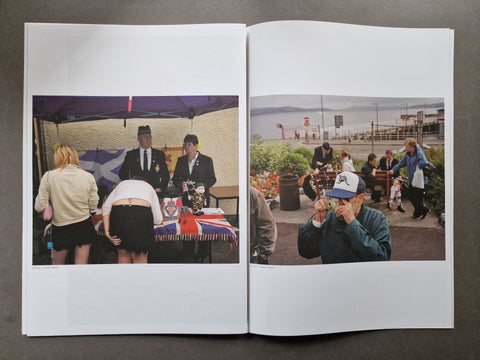 An open publication laid flat on a dark surface, showing two printed photographs side by side. The left page contains a photo of people gathered under a canopy around a table with a Union Jack cloth.